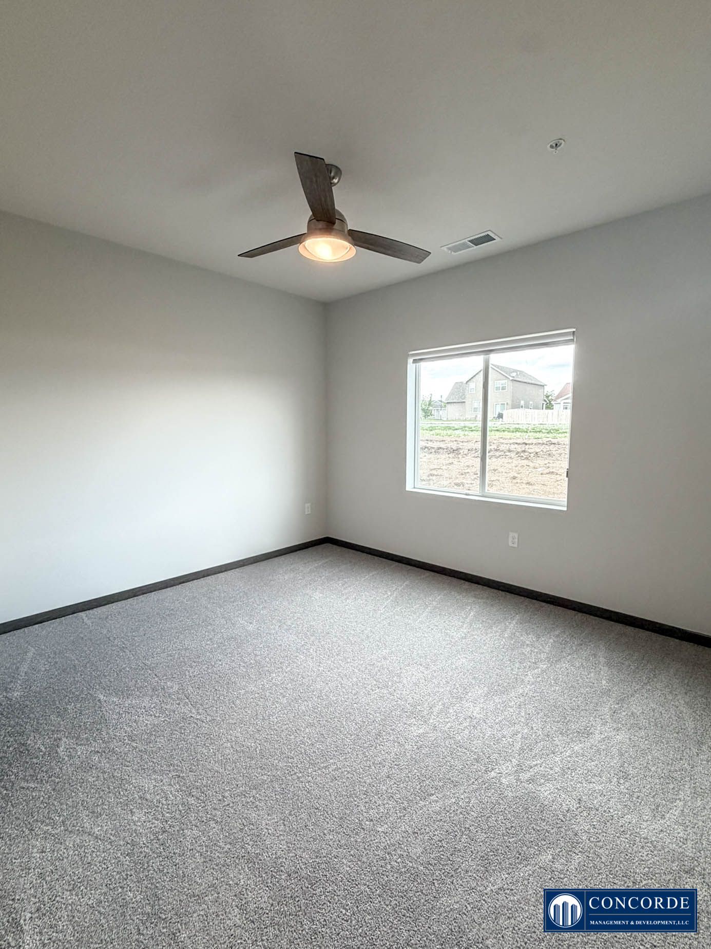 Empty white bedroom with gray carpet, ceiling fan, and a window overlooking a yard