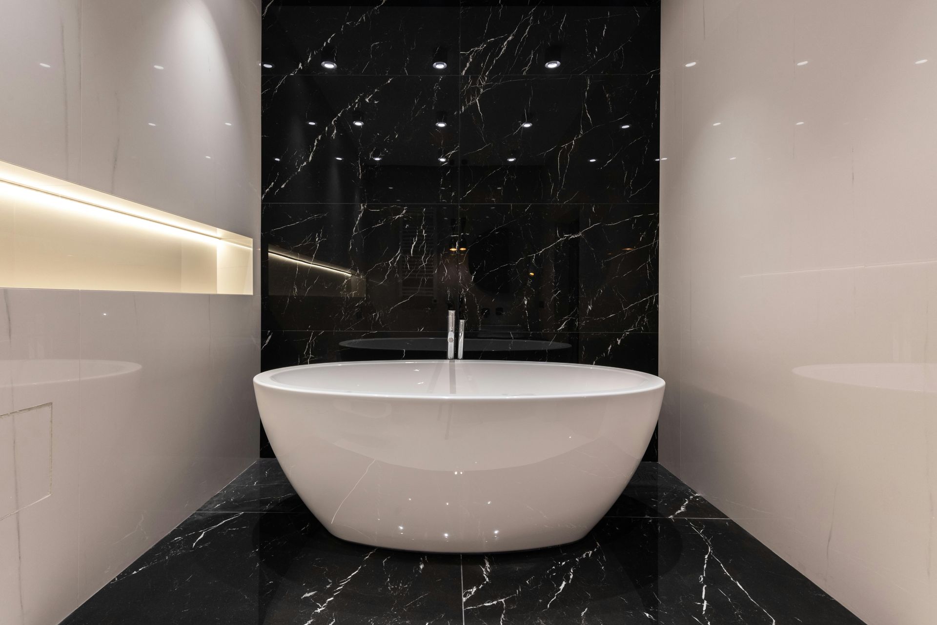 Modern bathroom with a white freestanding bathtub, black marble wall, and soft shelf lighting