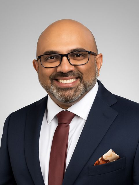 Man in navy suit, glasses, smiling, maroon tie, pocket square, against a gray backdrop.