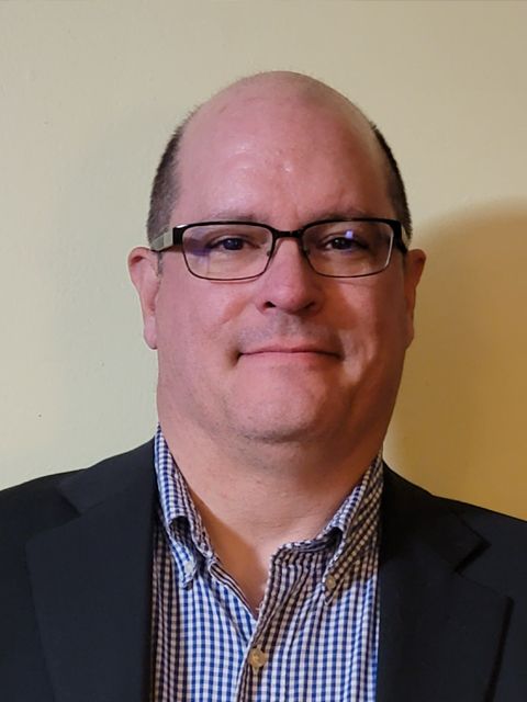 Man with glasses wearing a blazer and blue patterned shirt, against a pale yellow wall.