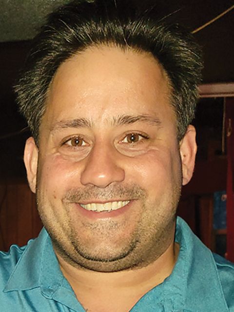 Man smiling, wearing a turquoise shirt, indoors with a dark background.