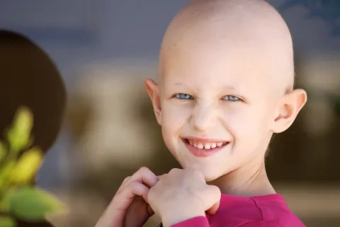 Smiling child with a shaved head wearing a pink shirt, hands under chin, outdoors.