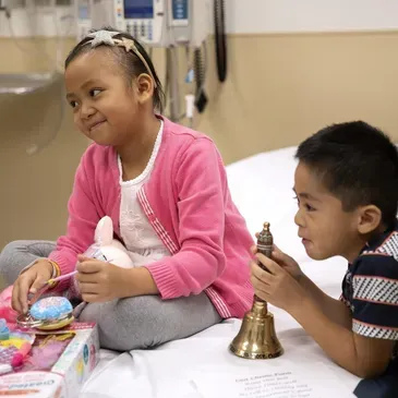 Two children in a hospital room: one with a pink cardigan looks at a toy; the other holds a bell.