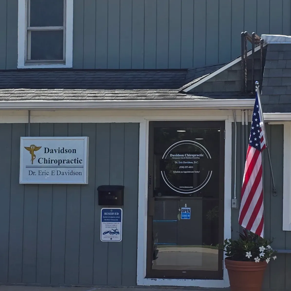 Gray building entrance with a sign, American flag, and a window beside a door with a wreath