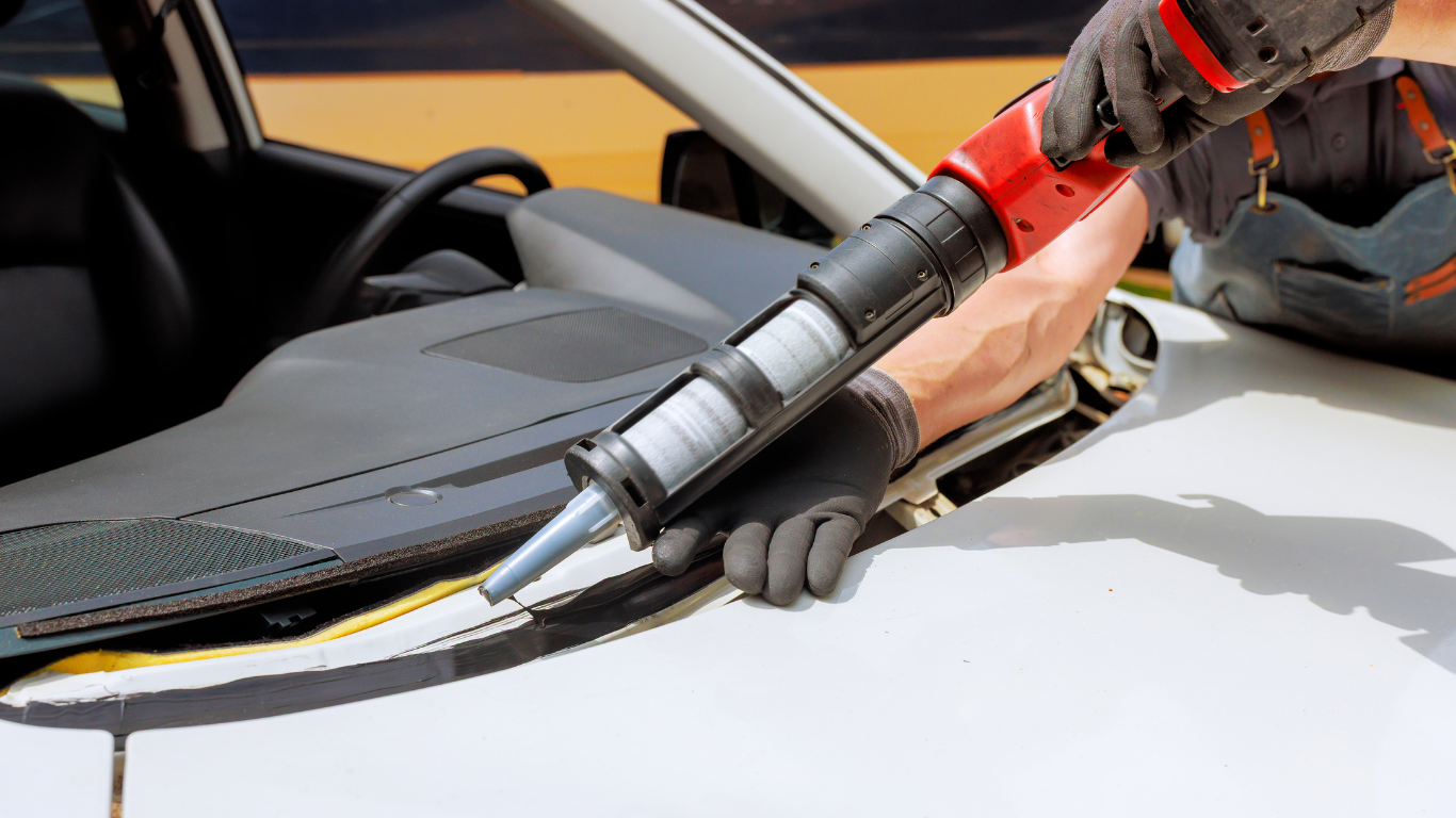 Person applying sealant around a car windshield with a red caulking gun.