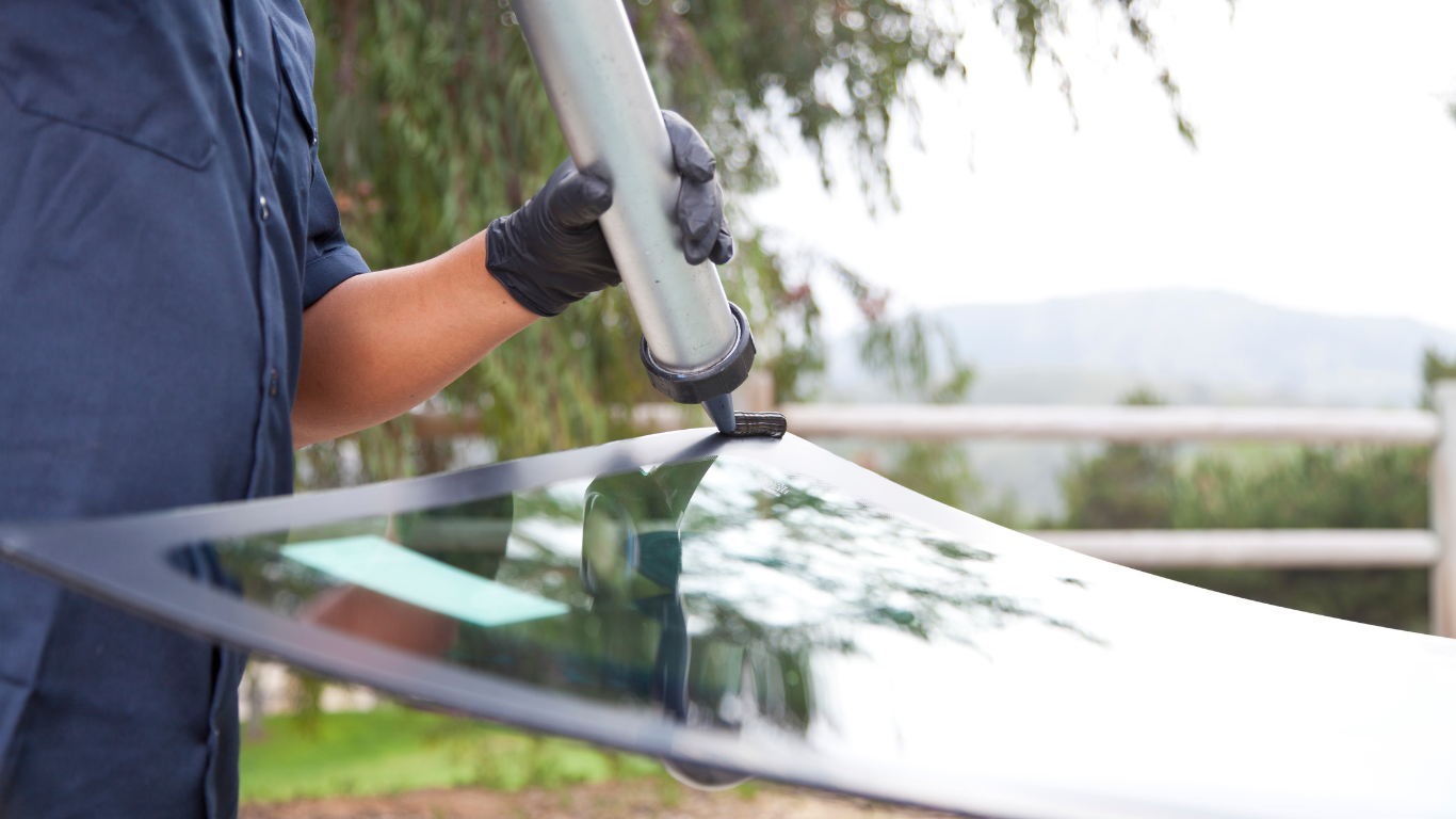 Person in blue shirt and black gloves applying sealant to a car windshield with a caulking gun outdoors.