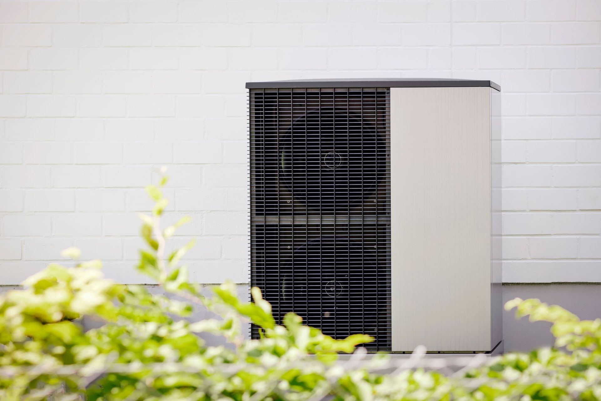 Outdoor HVAC unit with black grille and white side panel against a white wall, with blurred greenery in front