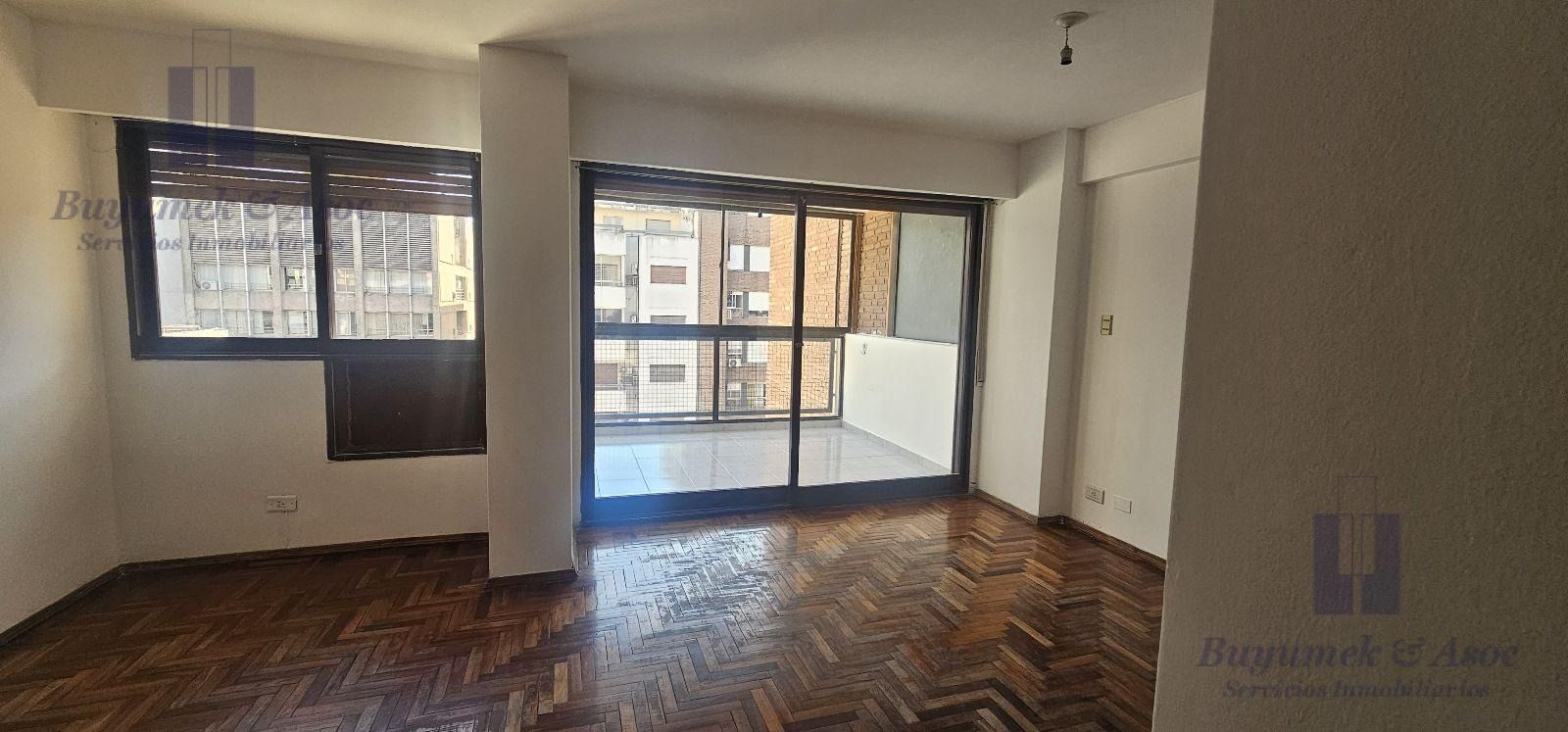 Salón vacío sin amueblar con suelo de parqué, grandes ventanales y puertas correderas de cristal que dan a un balcón.