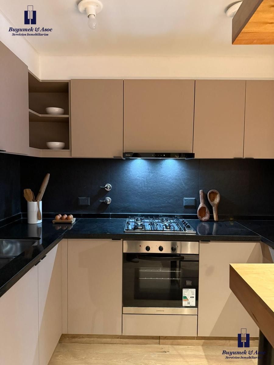 Apartamento moderno con zona de comedor y cocina, mesa de madera, isla negra y vistas a la ciudad a través de las ventanas.