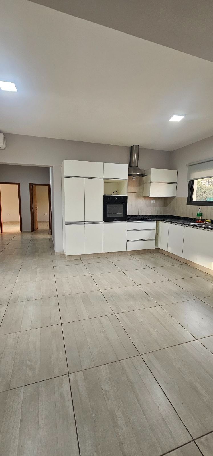 Cocina moderna vacía con gabinetes blancos, encimeras negras y piso de baldosas claras bajo iluminación empotrada.