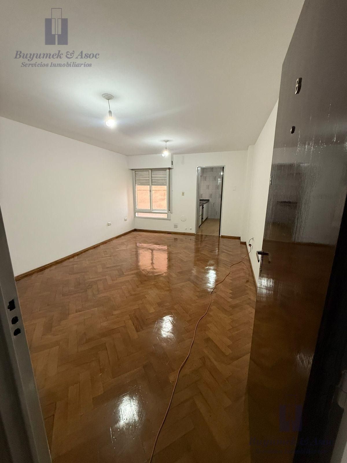 Habitación vacía con suelo de madera brillante, paredes blancas, luces en el techo y una ventana al fondo.