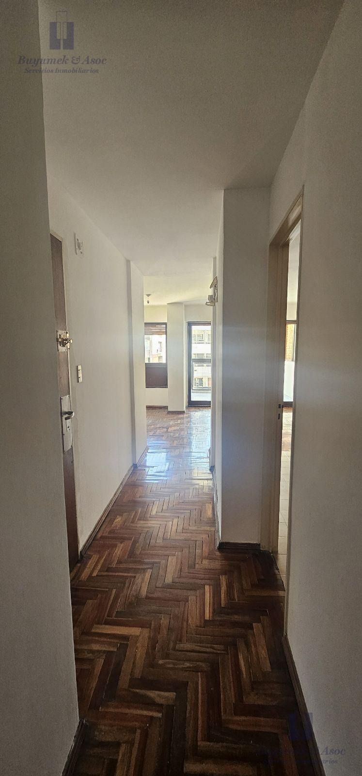 Salón vacío sin amueblar con suelo de parqué, grandes ventanales y puertas correderas de cristal que dan a un balcón.
