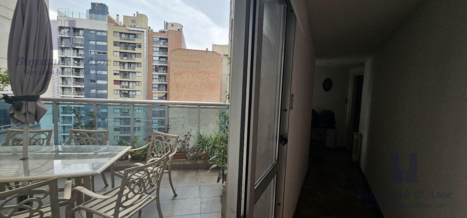 Balcón con mesa y sillas de exterior junto a un pasillo, con vistas a los edificios de apartamentos de la ciudad.