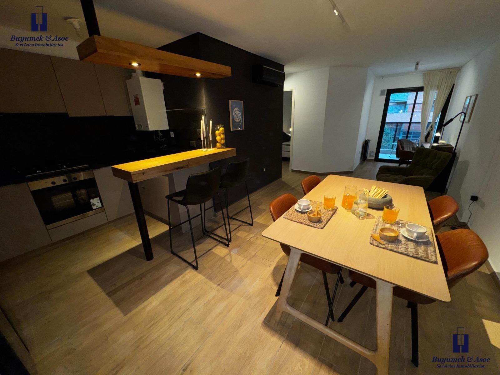 Apartamento moderno con zona de comedor y cocina, mesa de madera, isla negra y vistas a la ciudad a través de las ventanas.