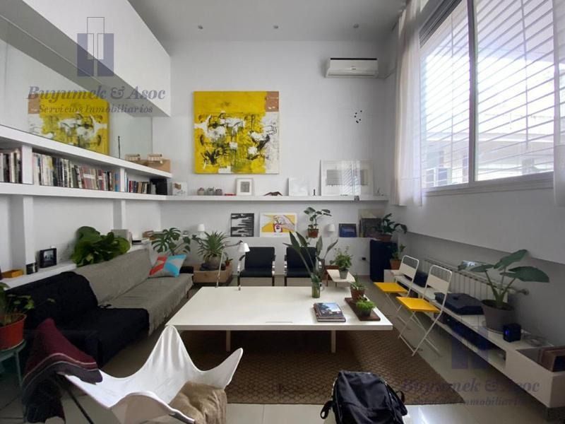 Acogedora sala de estar con estantes blancos, plantas, obras de arte amarillas y un sofá y una silla oscuros junto a la chimenea.