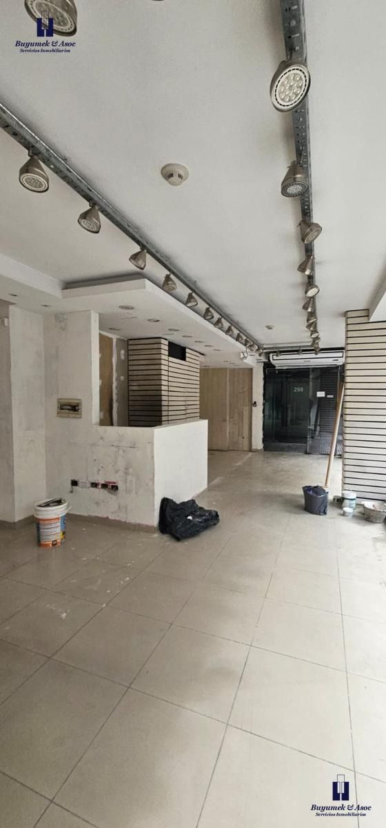 Pasillo comercial vacío con suelo de baldosas blancas, tuberías expuestas en el techo y cubos de limpieza cerca de una unidad de aire acondicionado montada en la pared.
