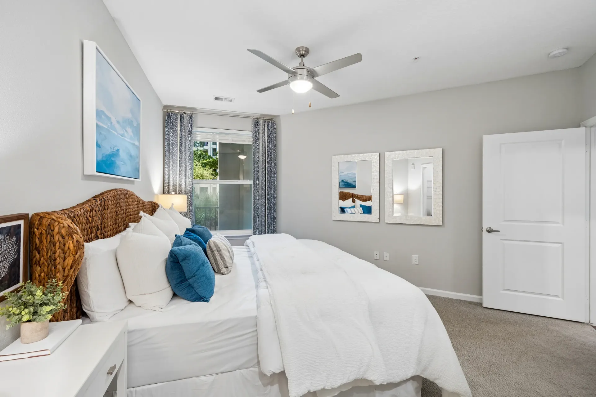 Cozy bedroom with a large wicker headboard and white bedding, featuring a ceiling fan and large windows at Atlantic on the Avenue in North Charleston, SC.