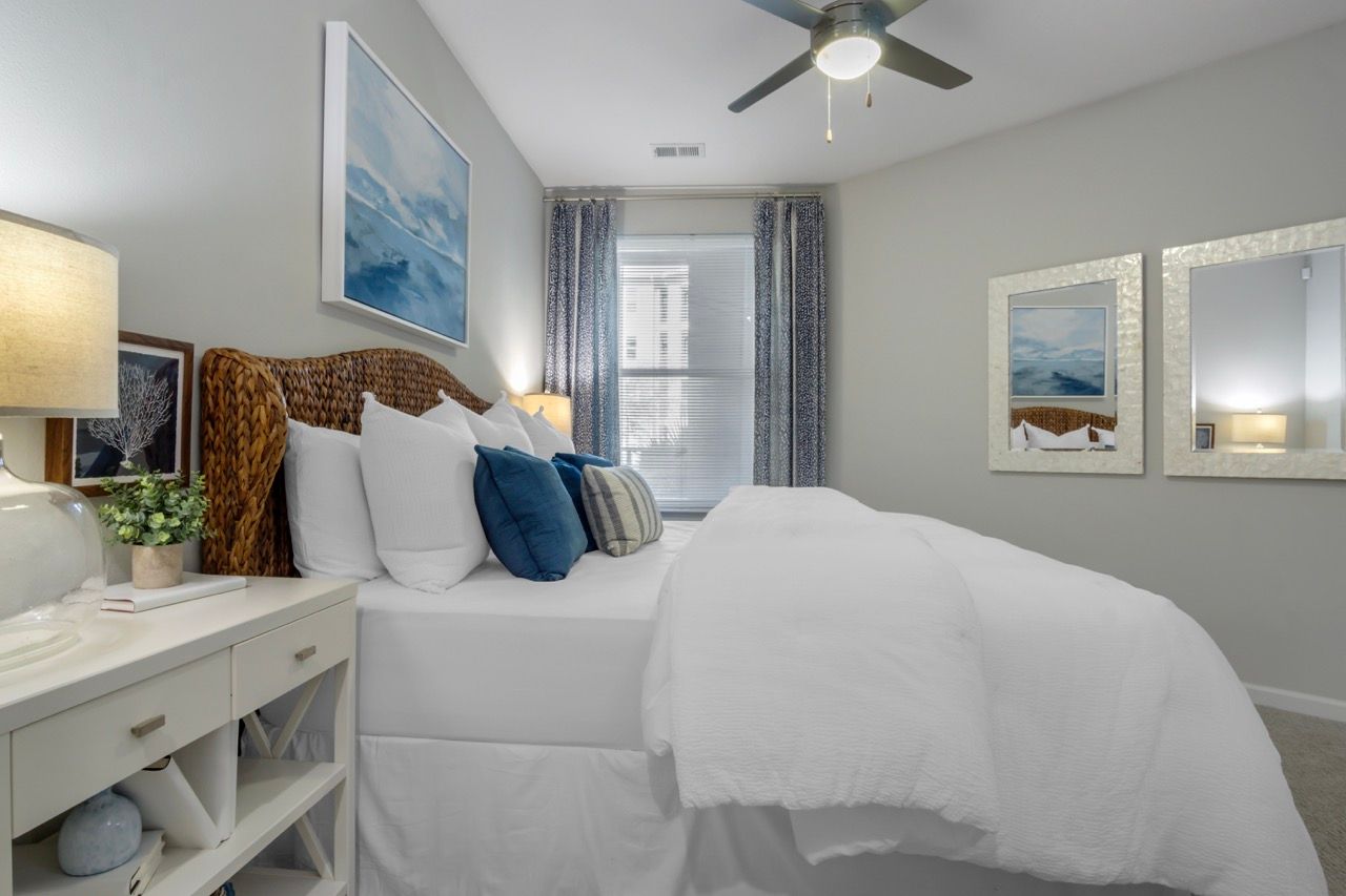 Bright modern apartment bedroom with white bedding, wicker headboard, blue accent pillows, and framed mirrors at Atlantic on the Avenue in North Charleston, SC.