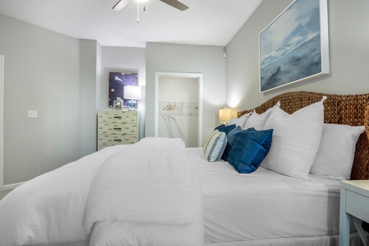 Bedroom in an apartment with a large white bed, wicker headboard, dresser, and open closet at Atlantic on the Avenue in North Charleston, SC.