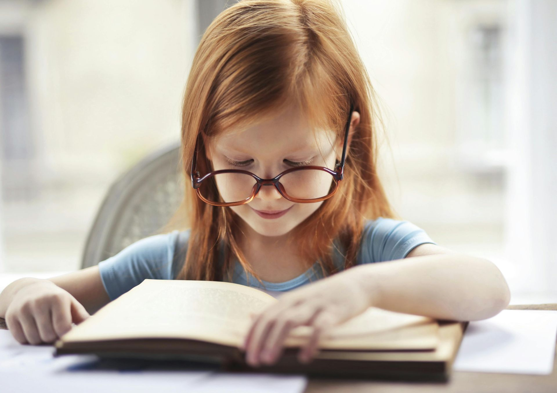 Child with red hair and glasses reading a book at a table by a bright window
