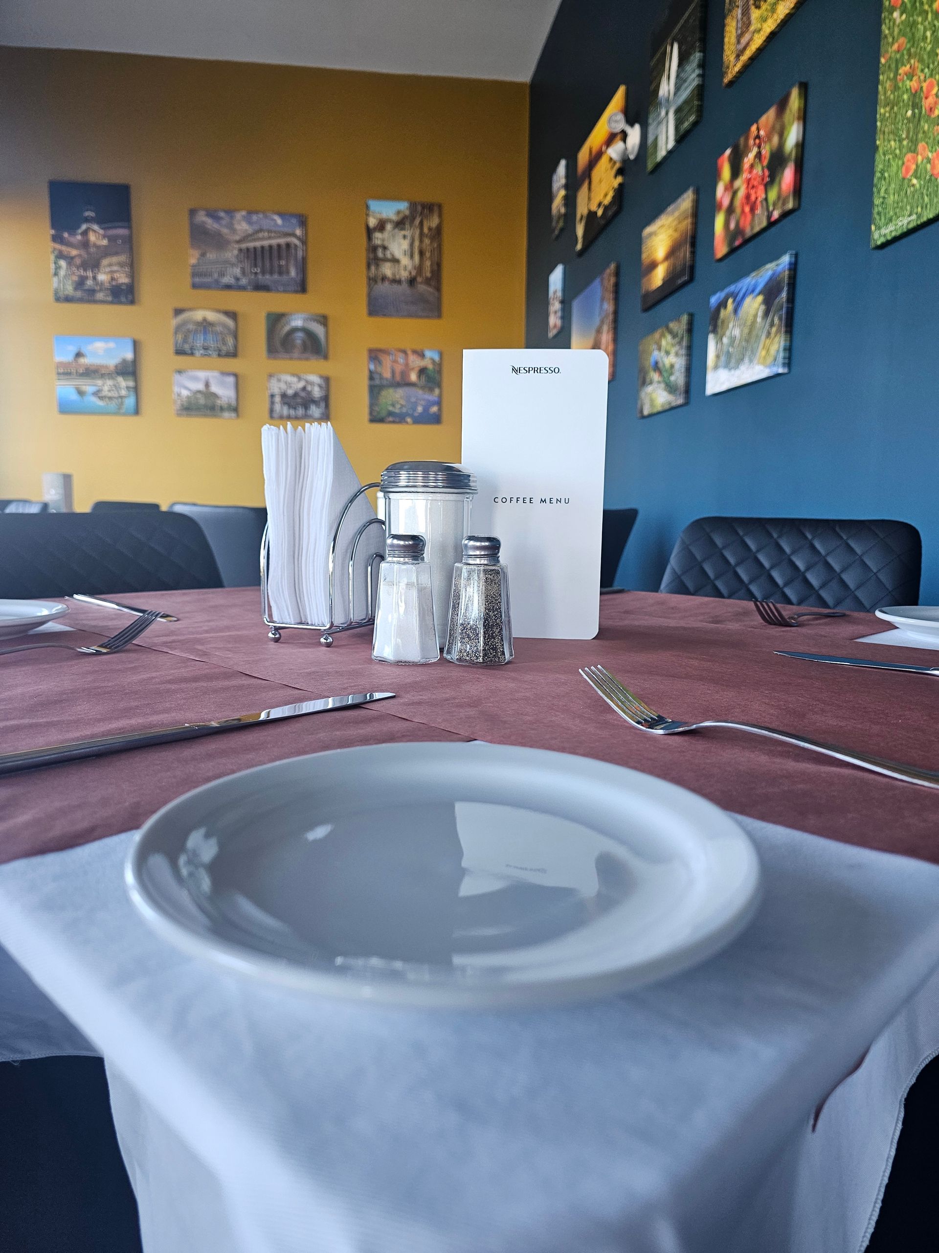 Dining table set with white plate and silverware; decorated with art on yellow and blue walls.