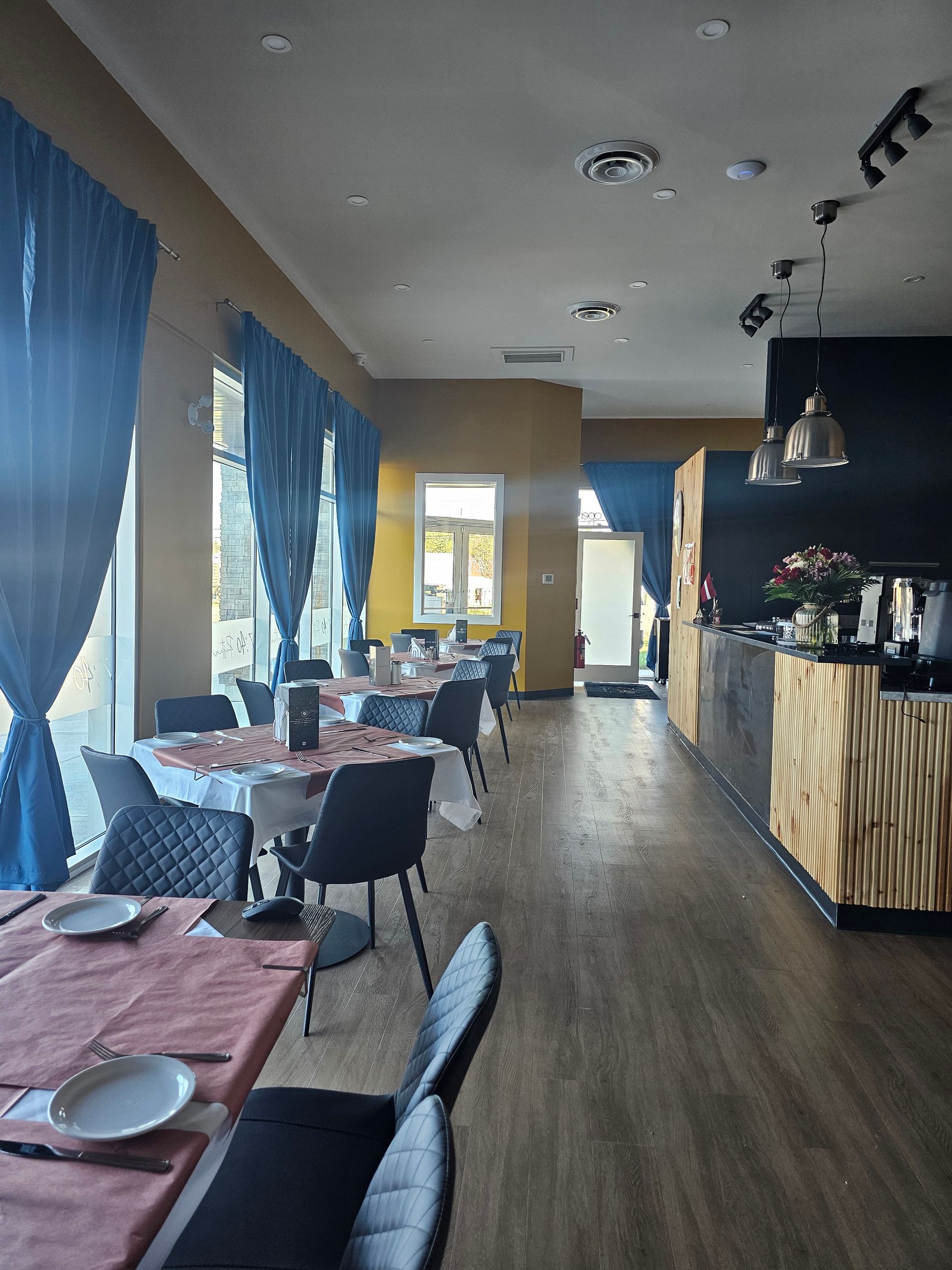 Interior of restaurant with tables set, blue curtains, wooden floor, and dark accent wall.