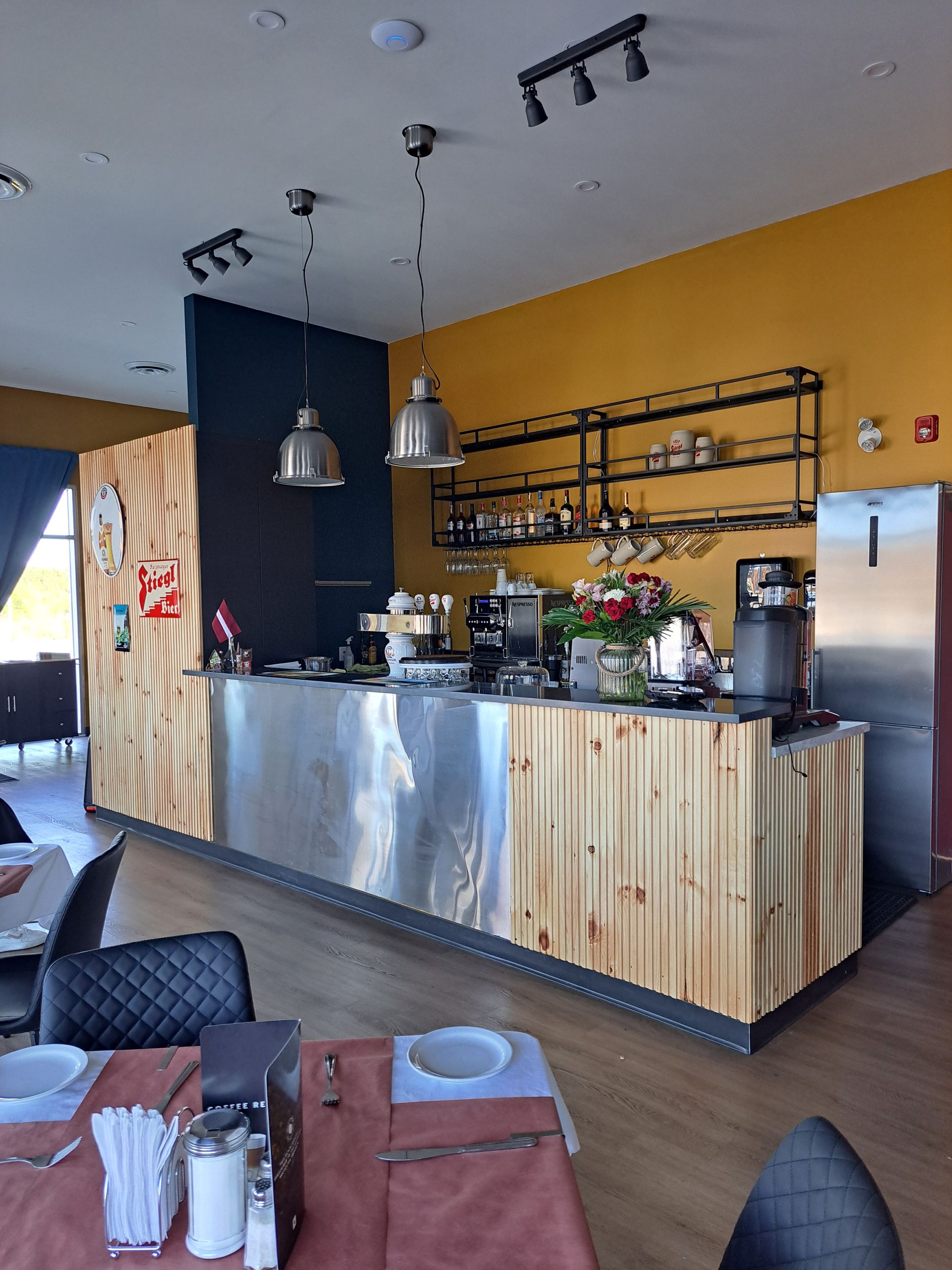 Interior of a cafe with a counter, tables, and a yellow wall with black shelves and lighting.