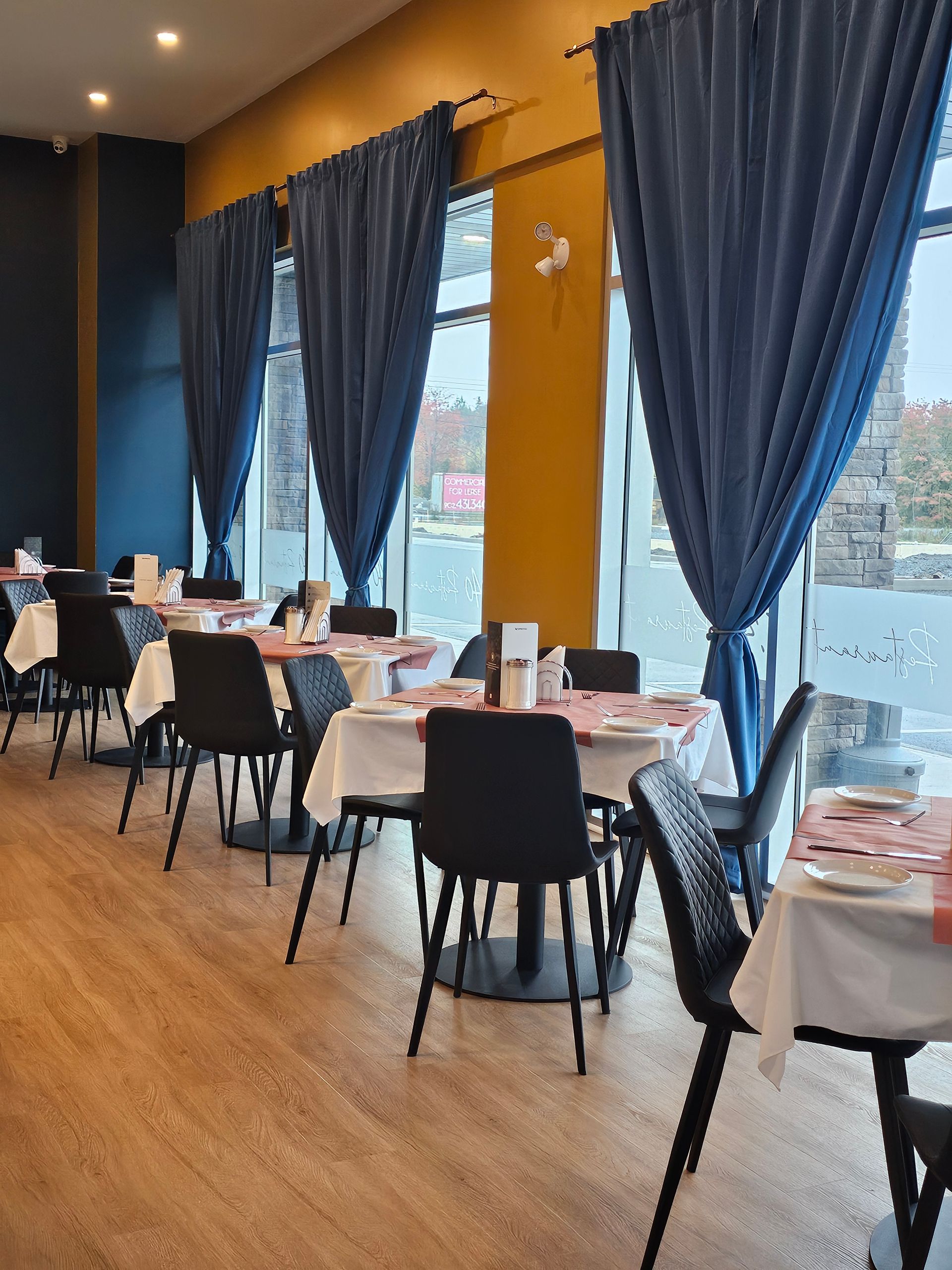Restaurant interior with tables set for dining, black chairs, blue curtains, and yellow walls.