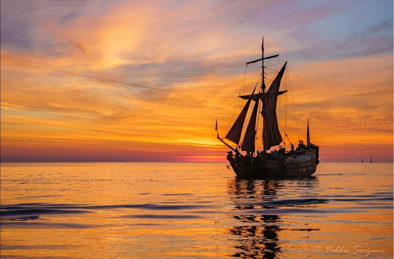 Sailboat silhouetted against a vibrant sunset sky and water reflecting the orange and gold hues.