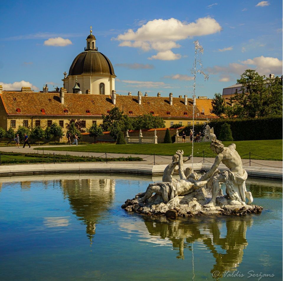 A baroque building with a dome reflected in a pond; white statue in foreground; blue sky.