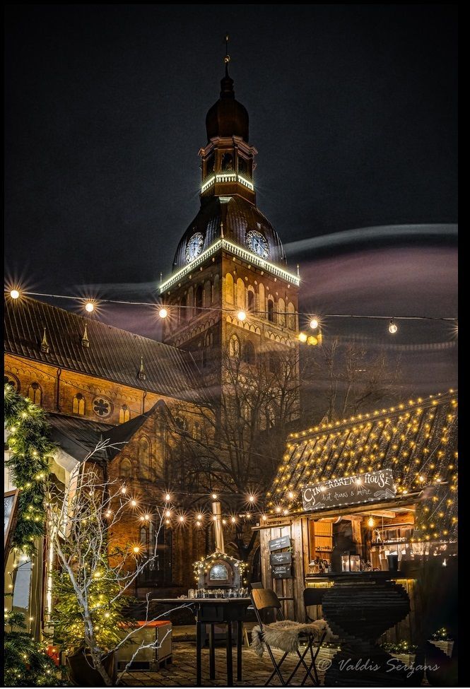 Night view of Riga Cathedral tower illuminated with holiday lights, surrounded by a festive market.