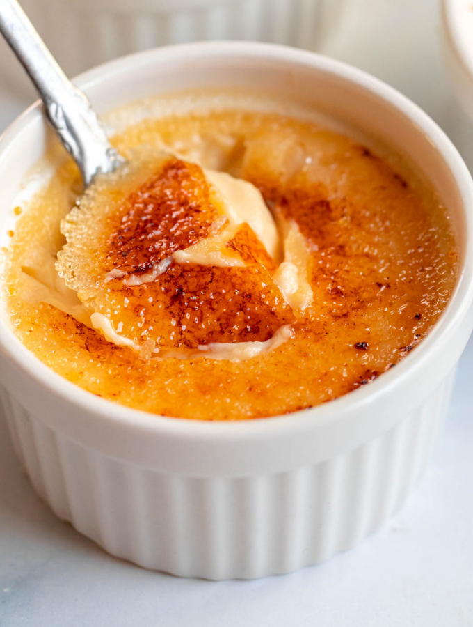 Crème brûlée in a white ramekin, with a spoon digging into the caramelized sugar crust.