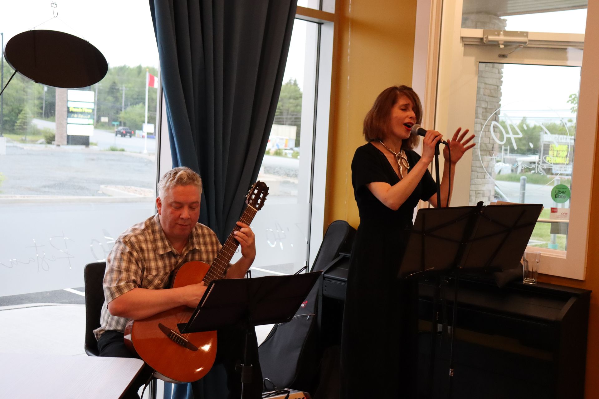 A man plays guitar and a woman sings into a microphone inside near a window.