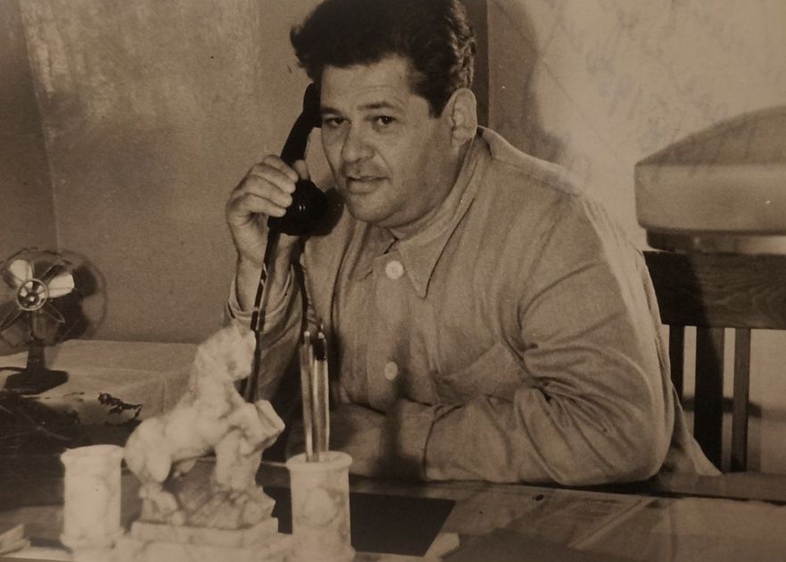 Man in light-colored jacket on the phone, seated at a desk with a decorative horse statue, desk lamp, and a fan.