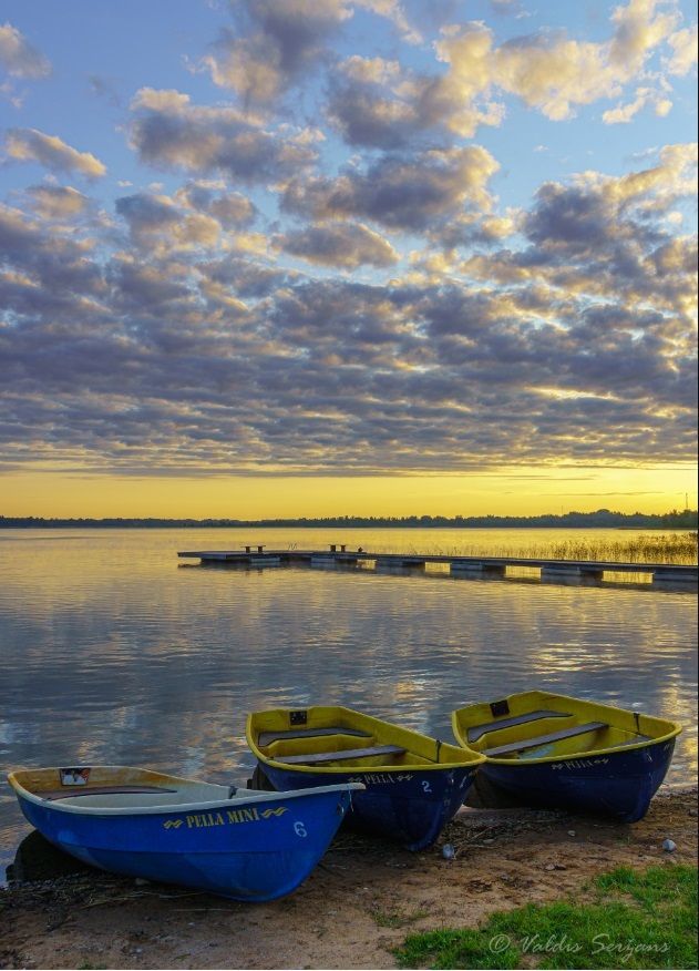 Three blue and yellow rowboats on a sandy shore under a cloudy sky reflecting in calm water.