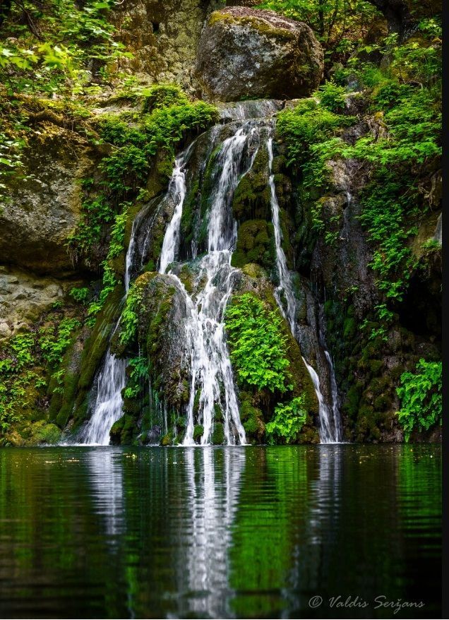 Waterfall cascading into a dark pond, surrounded by lush green foliage and rocky cliffs.