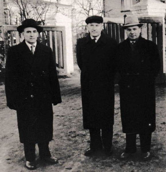 Three men in dark coats and hats stand outside, possibly near a gate or building.