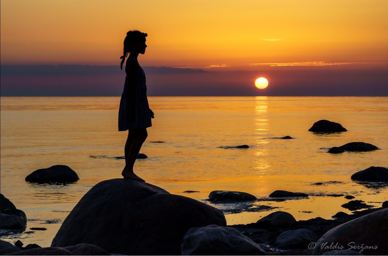Woman silhouetted on a rock, facing a sunset over the water; golden light.