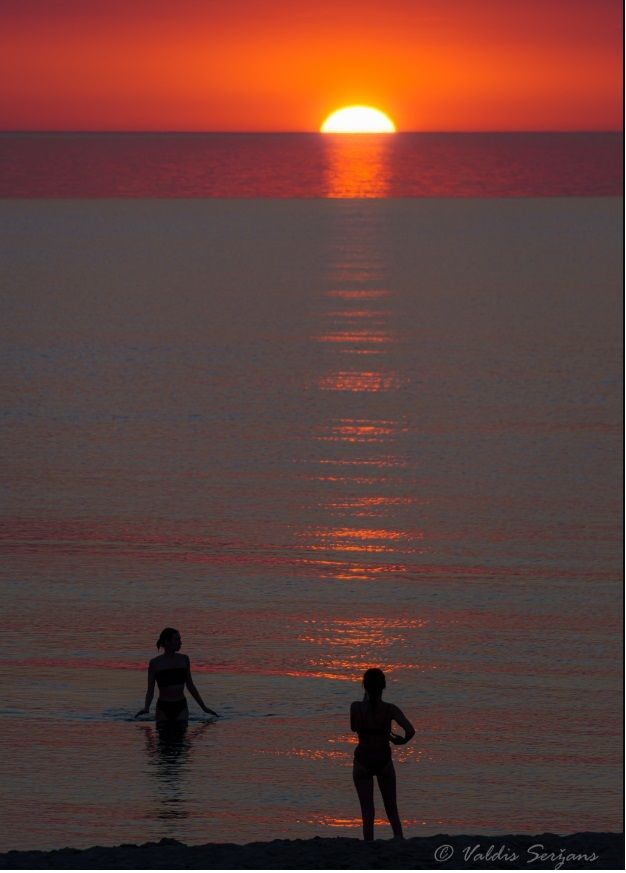 Two people in water watching a sunset over the sea; orange and red hues.
