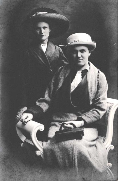 Two women in formal attire; one seated, holding a bag, the other standing behind. Black and white photo.