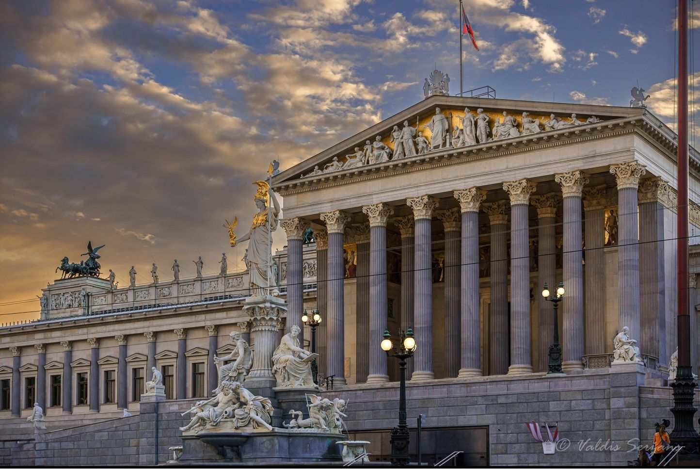 Austrian Parliament building with columns, sculptures, and flag, under a cloudy sunset sky.