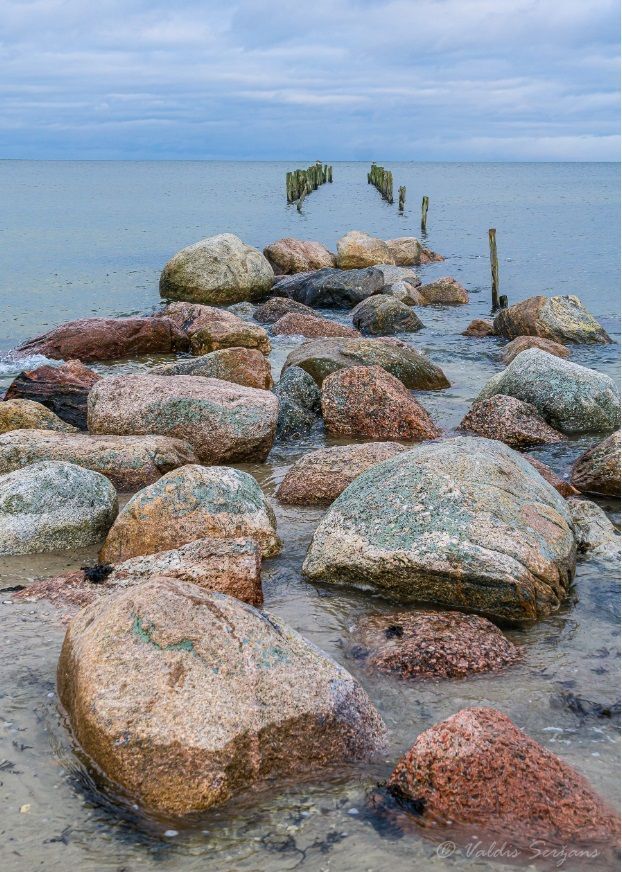 Rocks and wooden posts form a path into the sea under a cloudy sky.