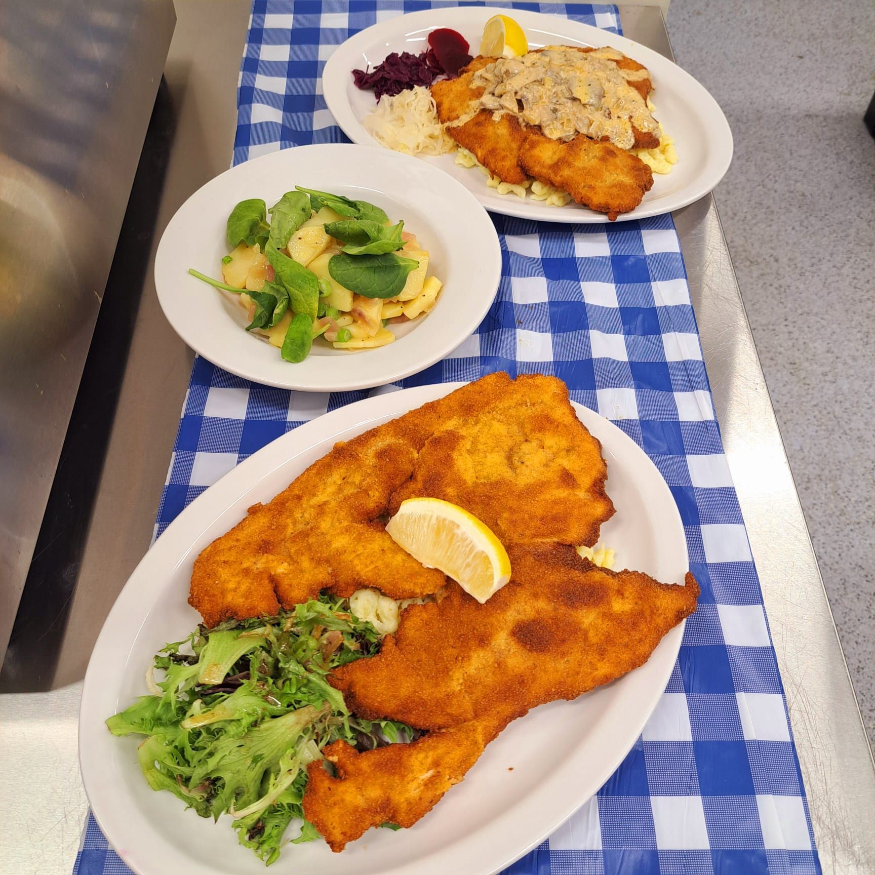 Platters of breaded schnitzel, potato salad, and salad on a blue checkered tablecloth.