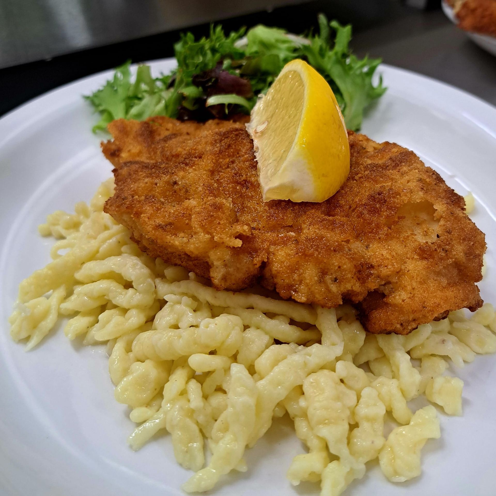 Breaded schnitzel with lemon wedge, served over spaetzle and greens on a white plate.