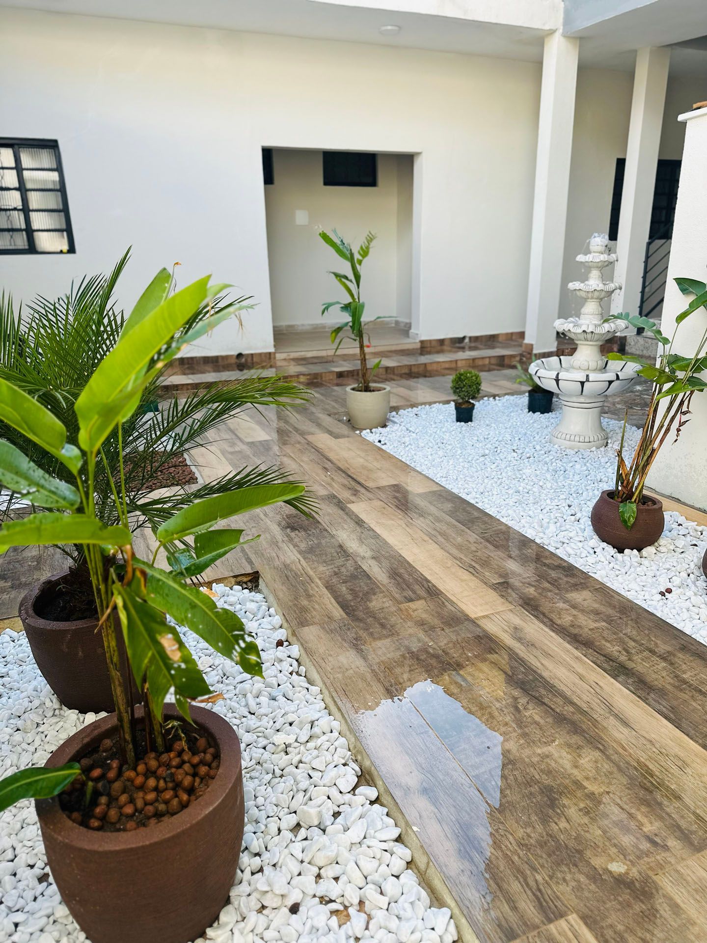 Pátio externo com caminho que imita madeira, seixos brancos, plantas em vasos e uma fonte contra paredes brancas.