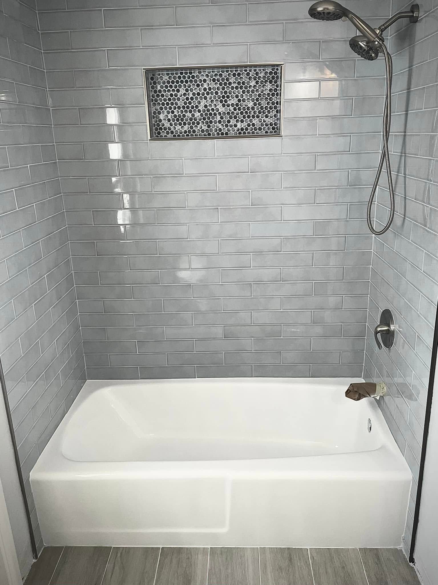 Bathtub in a gray-tiled shower stall, with a decorative alcove, and silver shower head and fixtures.