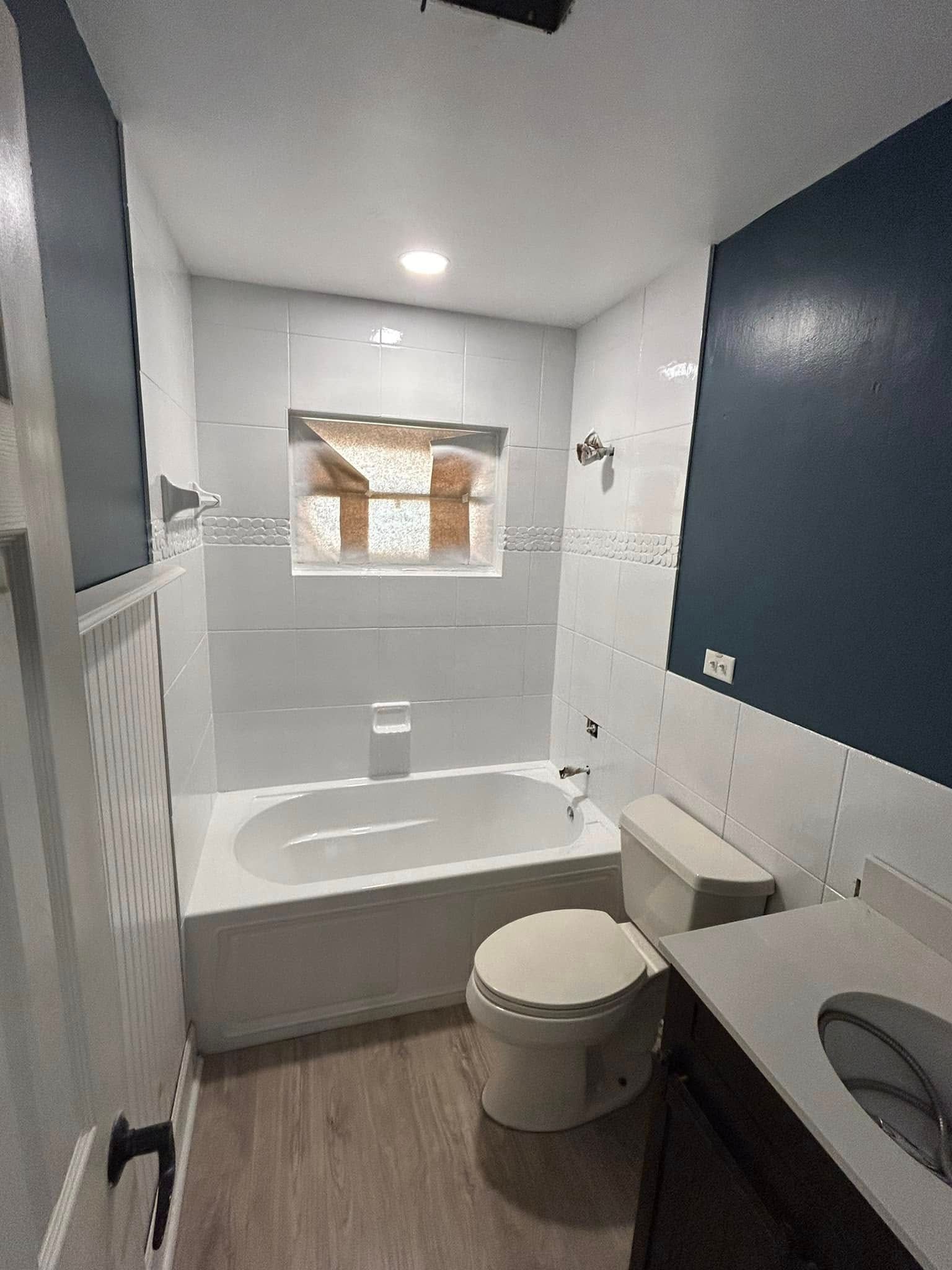 Bathroom with white tiled shower, navy wall, toilet, vanity, and wooden-look flooring.