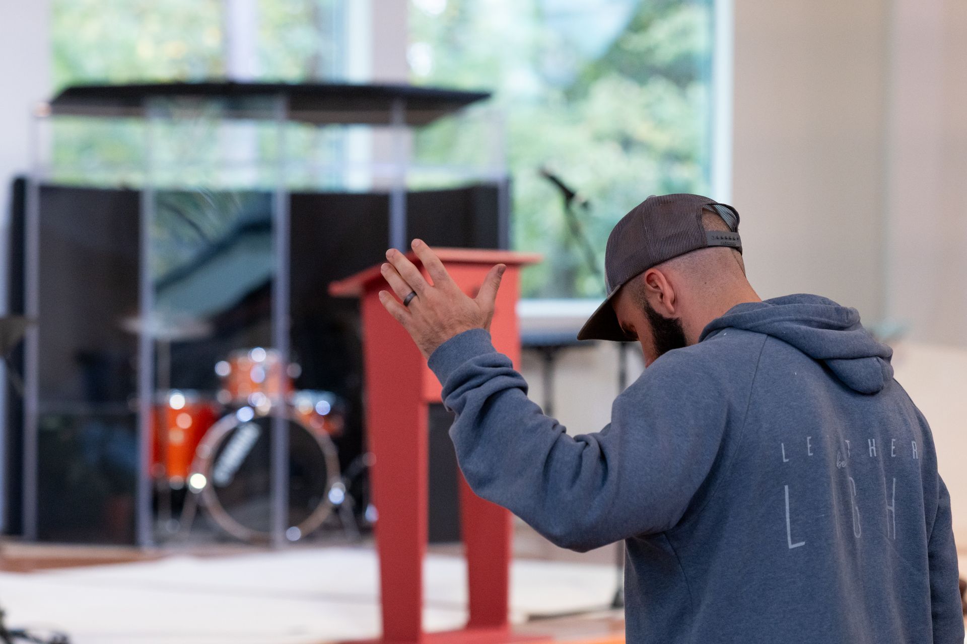 Man in a dark hoodie worshipping in front of glass-walled building and red drum set props