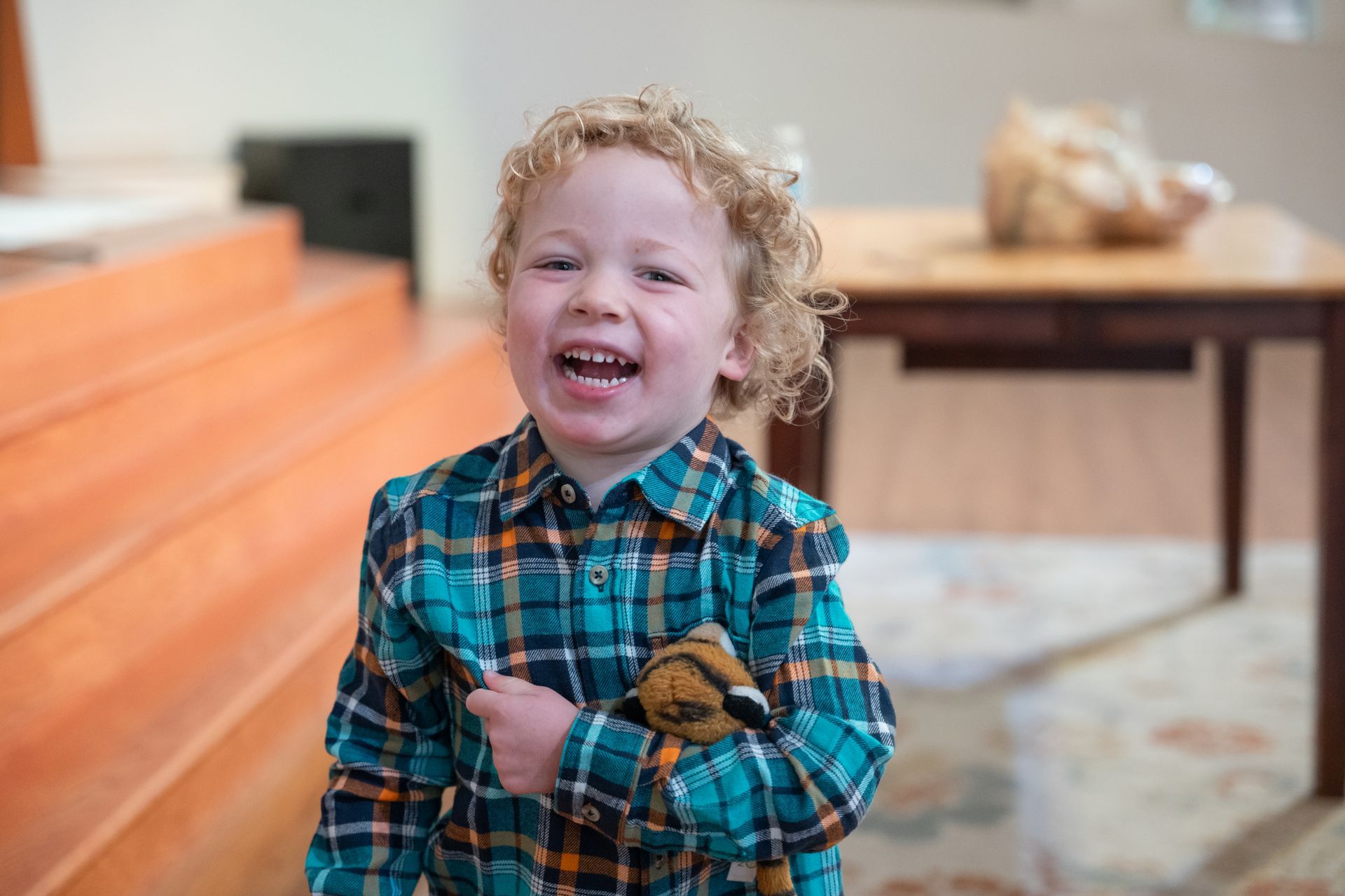 Smiling child in a teal plaid shirt standing indoors near orange steps and a table with a stuffed animal