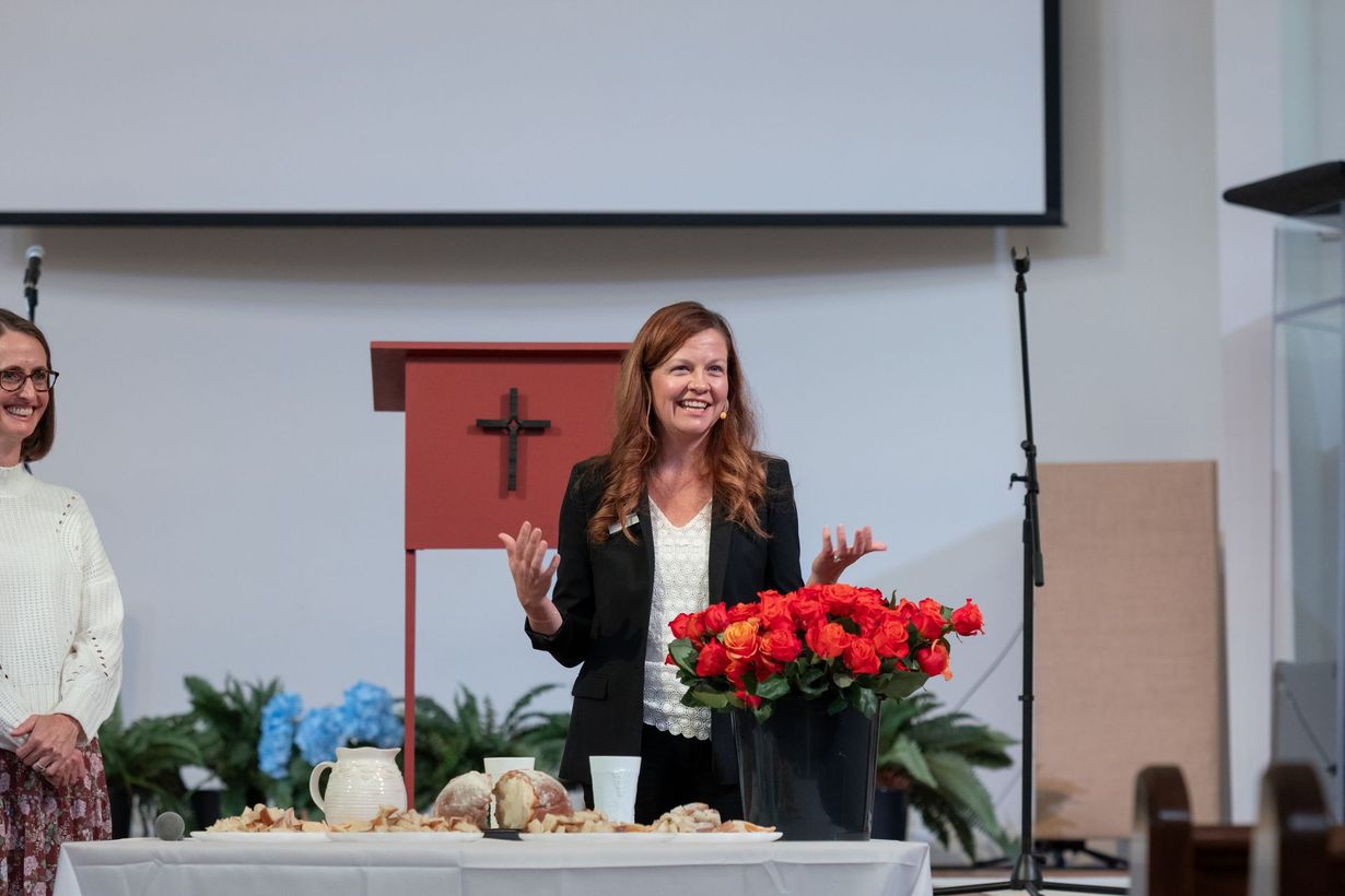 Woman speaking at a church podium beside a table with flowers and bread during a service